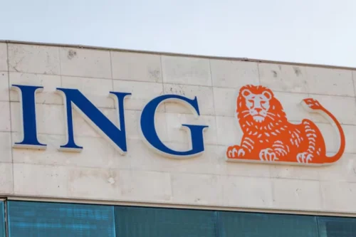 Foto em plano fechado da fachada de um edifício exibindo o logotipo da instituição financeira ING. À esquerda, destacam-se as letras "ING" em caixa alta, com uma fonte serifada na cor azul-escura e bordas brancas em relevo. À direita do texto, há o símbolo da marca: a ilustração estilizada de um leão deitado na cor laranja vibrante, também com contorno branco em relevo. O letreiro está fixado em uma parede de placas de concreto ou pedra clara, e logo abaixo é possível ver o topo de uma janela com vidros reflexivos azulados.