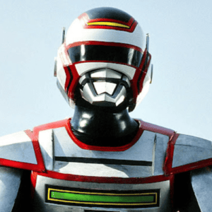 Fotografia em plano médio do herói japonês Jaspion, vestindo sua armadura metálica prateada com detalhes em vermelho e preto. Ele usa um capacete branco, vermelho e preto com visor escuro e detalhes prateados na região da boca. O peitoral da armadura possui uma faixa horizontal preta e um retângulo verde brilhante na parte inferior. O fundo é um céu claro e ensolarado.