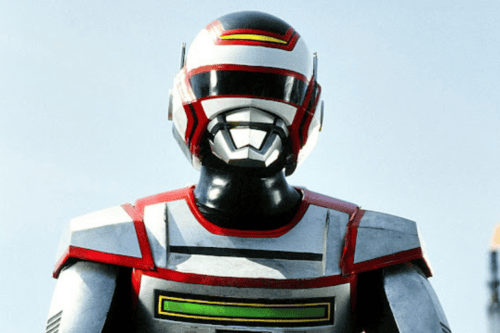 Fotografia em plano médio do herói japonês Jaspion, vestindo sua armadura metálica prateada com detalhes em vermelho e preto. Ele usa um capacete branco, vermelho e preto com visor escuro e detalhes prateados na região da boca. O peitoral da armadura possui uma faixa horizontal preta e um retângulo verde brilhante na parte inferior. O fundo é um céu claro e ensolarado.
