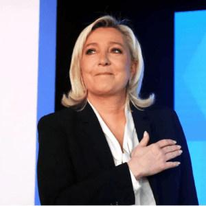 Fotografia de Marine Le Pen em plano médio, olhando ligeiramente para cima e para a esquerda com um leve sorriso. Ela tem cabelos loiros na altura dos ombros, usa um terno preto sobre uma blusa branca e está com a mão direita espalmada sobre o peito. O fundo é composto por painéis nas cores azul e branca.