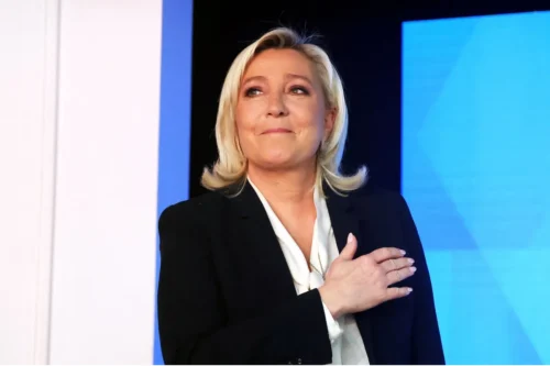 Fotografia de Marine Le Pen em plano médio, olhando ligeiramente para cima e para a esquerda com um leve sorriso. Ela tem cabelos loiros na altura dos ombros, usa um terno preto sobre uma blusa branca e está com a mão direita espalmada sobre o peito. O fundo é composto por painéis nas cores azul e branca.