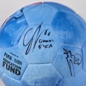 Uma imagem em plano médio e centralizada de uma bola de futebol azul-clara com textura aveludada. Na superfície da bola, há assinaturas feitas com caneta hidrográfica preta. A assinatura central destaca-se com o nome "Gianni" e a palavra "FIFA" escritos logo abaixo de uma rubrica estilizada. À direita, há outra assinatura com traços rápidos e um símbolo que se assemelha a uma coroa. No canto inferior esquerdo da bola, lê-se o texto estampado em letras pretas: "IN SUPPORT OF FIFA | GLOBAL CITIZEN EDUCATION FUND", disposto em três linhas, com a palavra "FUND" em tamanho maior e negrito. O fundo da imagem é cinza-claro e neutro.