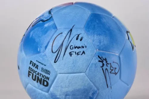 Uma imagem em plano médio e centralizada de uma bola de futebol azul-clara com textura aveludada. Na superfície da bola, há assinaturas feitas com caneta hidrográfica preta. A assinatura central destaca-se com o nome "Gianni" e a palavra "FIFA" escritos logo abaixo de uma rubrica estilizada. À direita, há outra assinatura com traços rápidos e um símbolo que se assemelha a uma coroa. No canto inferior esquerdo da bola, lê-se o texto estampado em letras pretas: "IN SUPPORT OF FIFA | GLOBAL CITIZEN EDUCATION FUND", disposto em três linhas, com a palavra "FUND" em tamanho maior e negrito. O fundo da imagem é cinza-claro e neutro.