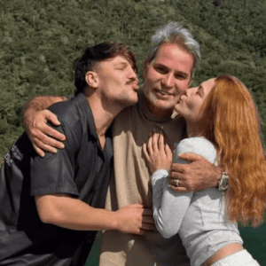 Três pessoas abraçadas em frente a uma paisagem natural de águas verdes e montanhas cobertas por vegetação densa. No centro, um homem de cabelos grisalhos e camiseta bege sorri para a câmera. À esquerda, um jovem homem de camisa preta o abraça pelos ombros e faz bico de beijo em direção à sua bochecha. À direita, uma jovem mulher ruiva, vestindo blusa cinza de manga comprida, também o abraça e faz bico de beijo em sua direção.