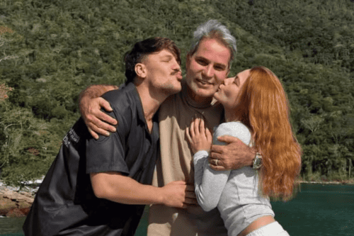 Três pessoas abraçadas em frente a uma paisagem natural de águas verdes e montanhas cobertas por vegetação densa. No centro, um homem de cabelos grisalhos e camiseta bege sorri para a câmera. À esquerda, um jovem homem de camisa preta o abraça pelos ombros e faz bico de beijo em direção à sua bochecha. À direita, uma jovem mulher ruiva, vestindo blusa cinza de manga comprida, também o abraça e faz bico de beijo em sua direção.