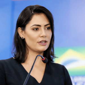Fotografia em plano médio de Michelle Bolsonaro. Ela é uma mulher de pele clara, cabelos pretos na altura dos ombros e olhos castanhos. Está levemente virada de perfil para a direita, falando ao microfone de haste preta que aparece em primeiro plano. Ela usa um vestido preto de decote em V e brincos pequenos escuros. O fundo é composto por uma parede azul e, de forma desfocada à direita, a bandeira do Brasil.