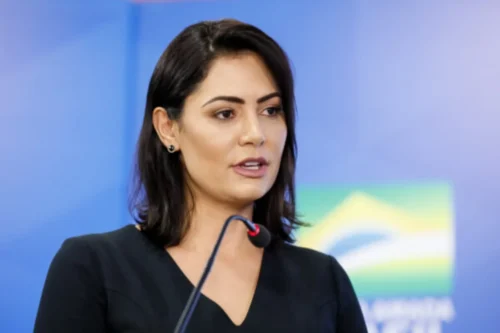 Fotografia em plano médio de Michelle Bolsonaro. Ela é uma mulher de pele clara, cabelos pretos na altura dos ombros e olhos castanhos. Está levemente virada de perfil para a direita, falando ao microfone de haste preta que aparece em primeiro plano. Ela usa um vestido preto de decote em V e brincos pequenos escuros. O fundo é composto por uma parede azul e, de forma desfocada à direita, a bandeira do Brasil.