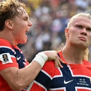 Dois jogadores de futebol da seleção da Noruega com camisas vermelhas e detalhes em azul e branco. À esquerda, um jogador com cabelo loiro encaracolado grita entusiasmado de perfil, segurando o ombro do companheiro de equipe. À direita, o jogador Erling Haaland, com o cabelo loiro preso, olha para a frente com uma expressão séria e focada. O fundo mostra uma arquibancada desfocada com público.