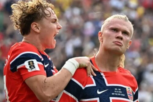 Dois jogadores de futebol da seleção da Noruega com camisas vermelhas e detalhes em azul e branco. À esquerda, um jogador com cabelo loiro encaracolado grita entusiasmado de perfil, segurando o ombro do companheiro de equipe. À direita, o jogador Erling Haaland, com o cabelo loiro preso, olha para a frente com uma expressão séria e focada. O fundo mostra uma arquibancada desfocada com público.