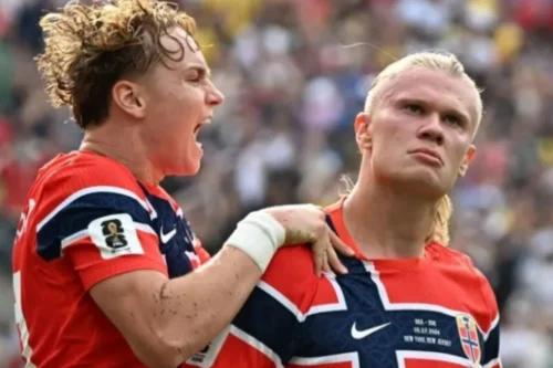 Dois jogadores de futebol da seleção da Noruega com camisas vermelhas e detalhes em azul e branco. À esquerda, um jogador com cabelo loiro encaracolado grita entusiasmado de perfil, segurando o ombro do companheiro de equipe. À direita, o jogador Erling Haaland, com o cabelo loiro preso, olha para a frente com uma expressão séria e focada. O fundo mostra uma arquibancada desfocada com público.