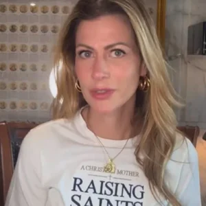 oto de Pietra Bertolazzi, uma mulher branca de cabelos loiros e ondulados. Ela veste uma camiseta branca com a estampa em inglês 'A Christian Mother Raising Saints', brincos de argola dourados e um colar com pingente religioso. Ao fundo, vê-se uma parede decorada com um quadro com selos ou ilustrações organizadas em colunas e uma luminária acesa.
