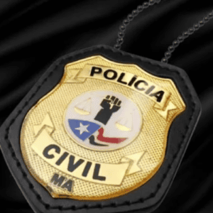 Um distintivo dourado da Polícia Civil do Estado do Maranhão (indicado pela sigla "MA" na parte inferior), fixado em um suporte de couro preto com uma corrente de metal ao fundo. No centro do distintivo metálico, há um brasão redondo com detalhes em azul, vermelho e branco, exibindo o desenho de um punho cerrado erguido, uma balança da justiça e a bandeira estilizada do estado. O distintivo está posicionado diagonalmente sobre um fundo de tecido preto acetinado e brilhante.