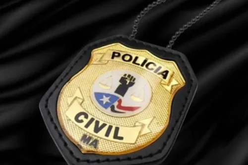 Um distintivo dourado da Polícia Civil do Estado do Maranhão (indicado pela sigla "MA" na parte inferior), fixado em um suporte de couro preto com uma corrente de metal ao fundo. No centro do distintivo metálico, há um brasão redondo com detalhes em azul, vermelho e branco, exibindo o desenho de um punho cerrado erguido, uma balança da justiça e a bandeira estilizada do estado. O distintivo está posicionado diagonalmente sobre um fundo de tecido preto acetinado e brilhante.
