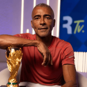 Homem de pele parda, meia-idade, com cabelos curtos e grisalhos, vestindo uma camiseta vermelha de manga curta. Ele está sentado em uma poltrona branca, com o braço direito apoiado sobre o topo do troféu da Copa do Mundo da FIFA. Ao fundo, há um estúdio iluminado com luzes verticais brancas e um painel azul com a inscrição "R Tv.".