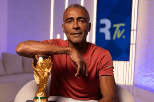 Homem de pele parda, meia-idade, com cabelos curtos e grisalhos, vestindo uma camiseta vermelha de manga curta. Ele está sentado em uma poltrona branca, com o braço direito apoiado sobre o topo do troféu da Copa do Mundo da FIFA. Ao fundo, há um estúdio iluminado com luzes verticais brancas e um painel azul com a inscrição "R Tv.".