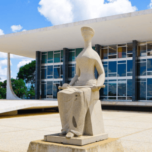 Fotografia da fachada do Supremo Tribunal Federal (STF) em Brasília, sob um céu azul com nuvens brancas. Em primeiro plano, centralizada, destaca-se a escultura de granito "A Justiça", de Alfredo Ceschiatti, que retrata uma mulher sentada, de olhos vendados, segurando uma espada sobre o colo. Ao fundo, ergue-se o edifício de arquitetura moderna projetado por Oscar Niemeyer, caracterizado por suas colunas brancas curvas e uma extensa fachada de vidro que reflete o ambiente externo.