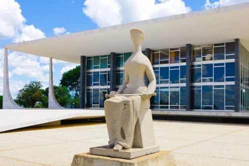 Fotografia da fachada do Supremo Tribunal Federal (STF) em Brasília, sob um céu azul com nuvens brancas. Em primeiro plano, centralizada, destaca-se a escultura de granito "A Justiça", de Alfredo Ceschiatti, que retrata uma mulher sentada, de olhos vendados, segurando uma espada sobre o colo. Ao fundo, ergue-se o edifício de arquitetura moderna projetado por Oscar Niemeyer, caracterizado por suas colunas brancas curvas e uma extensa fachada de vidro que reflete o ambiente externo.