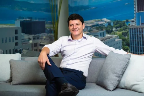Homem de cabelo curto e escuro, sorrindo, sentado confortavelmente em um sofá cinza com almofadas. Ele veste uma camisa de botão clara com as mangas dobradas e calça social escura. Ao fundo, há uma grande janela ou painel mostrando uma paisagem urbana com prédios e o mar ao fundo.