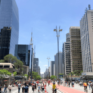 Uma vista ampla da Avenida Paulista, em São Paulo, em um dia ensolarado com céu azul claro. A avenida está fechada para carros e repleta de pedestres caminhando e ciclistas andando por uma ciclofaixa vermelha central. Ambos os lados da via são ladeados por grandes edifícios comerciais modernos, incluindo arranha-céus espelhados e de concreto, além de antenas de transmissão ao fundo. À direita, destaca-se a arquitetura marcante em formato de pirâmide do prédio do FIESP.