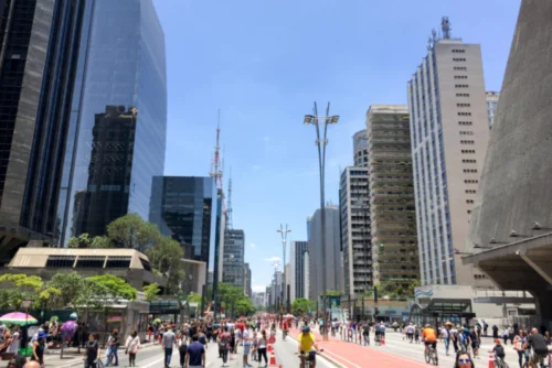 Uma vista ampla da Avenida Paulista, em São Paulo, em um dia ensolarado com céu azul claro. A avenida está fechada para carros e repleta de pedestres caminhando e ciclistas andando por uma ciclofaixa vermelha central. Ambos os lados da via são ladeados por grandes edifícios comerciais modernos, incluindo arranha-céus espelhados e de concreto, além de antenas de transmissão ao fundo. À direita, destaca-se a arquitetura marcante em formato de pirâmide do prédio do FIESP.