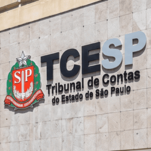 Fachada de placas de pedra clara com o letreiro do "TCESP - Tribunal de Contas do Estado de São Paulo" fixado na parede. À esquerda do texto em letras pretas e cinzas, há o brasão do Estado de São Paulo colorido em vermelho, verde e prata. A foto foi tirada em um ângulo ligeiramente inclinado de baixo para cima.
