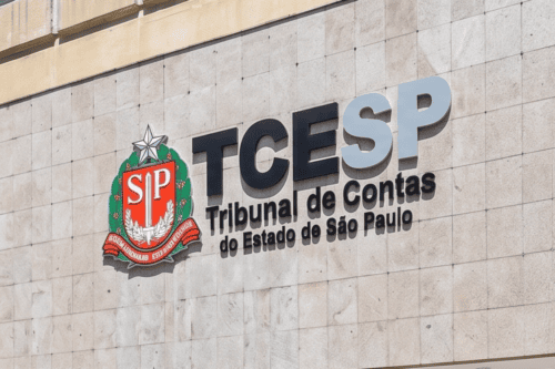 Fachada de placas de pedra clara com o letreiro do "TCESP - Tribunal de Contas do Estado de São Paulo" fixado na parede. À esquerda do texto em letras pretas e cinzas, há o brasão do Estado de São Paulo colorido em vermelho, verde e prata. A foto foi tirada em um ângulo ligeiramente inclinado de baixo para cima.