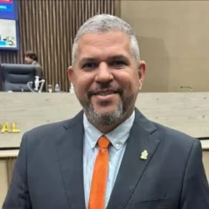 Homem de pele clara, cabelos curtos grisalhos e barba por fazer, sorrindo para a foto. Ele veste um terno preto, camisa social clara e uma gravata laranja vibrante. Ao fundo, há um ambiente institucional com painéis de madeira, cadeiras de escritório pretas e telas de monitor, característico de um plenário ou câmara municipal. Na bancada atrás dele, lê-se parcialmente a inscrição "MUNICIPAL NAUS".