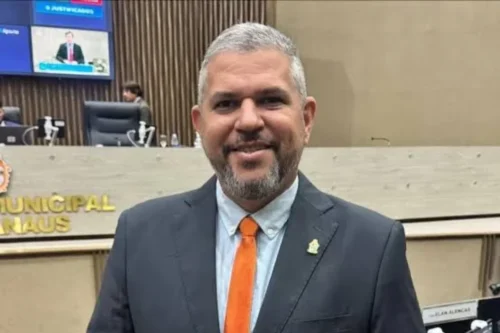 Homem de pele clara, cabelos curtos grisalhos e barba por fazer, sorrindo para a foto. Ele veste um terno preto, camisa social clara e uma gravata laranja vibrante. Ao fundo, há um ambiente institucional com painéis de madeira, cadeiras de escritório pretas e telas de monitor, característico de um plenário ou câmara municipal. Na bancada atrás dele, lê-se parcialmente a inscrição "MUNICIPAL NAUS".