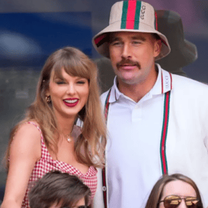 Fotografia em plano médio da cantora Taylor Swift e do jogador de futebol americano Travis Kelce, sorridentes, em um evento público. Taylor está à esquerda, com cabelos loiros e franja, usando batom vermelho marcante e um vestido com estampa xadrez miúda em tons de vermelho e branco. Travis está ao seu lado, à direita, com um bigode proeminente, vestindo uma camisa polo branca com detalhes em listras verdes e vermelhas, além de um chapéu estilo bucket de cor bege com as mesmas listras coloridas. O fundo sugere uma área de camarote ou arquibancada.