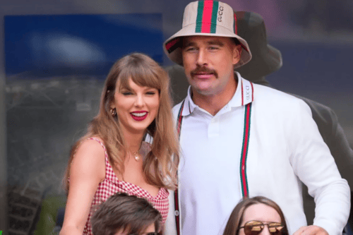 Fotografia em plano médio da cantora Taylor Swift e do jogador de futebol americano Travis Kelce, sorridentes, em um evento público. Taylor está à esquerda, com cabelos loiros e franja, usando batom vermelho marcante e um vestido com estampa xadrez miúda em tons de vermelho e branco. Travis está ao seu lado, à direita, com um bigode proeminente, vestindo uma camisa polo branca com detalhes em listras verdes e vermelhas, além de um chapéu estilo bucket de cor bege com as mesmas listras coloridas. O fundo sugere uma área de camarote ou arquibancada.