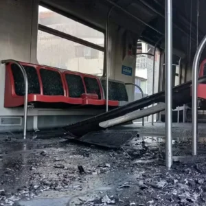 Fotografia do interior de um vagão de trem urbano danificado após um incêndio ou vandalismo. O chão está coberto de fuligem, cinzas e escombros escuros. Uma parte do teto ou estrutura interna caiu, ficando inclinada entre as fileiras de assentos vermelhos. Pelo vidro da janela e pelas portas abertas ao fundo, avistam-se edificações da cidade.