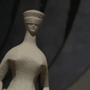 Fotografia em plano médio focada na parte superior da escultura "A Justiça", do artista Alfredo Ceschiatti, localizada em Brasília. A estátua de granito cinza-claro representa uma mulher estilizada com os olhos vendados, expressando neutralidade e imparcialidade. O fundo está escuro e desfocado, destacando a textura áspera da escultura sob uma iluminação suave que vem da esquerda.