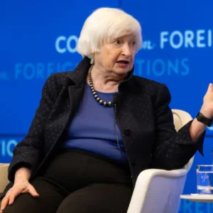 Janet Yellen, uma mulher idosa de cabelos brancos curtos com franja, está sentada em uma poltrona branca falando em um evento. Ela veste uma blusa azul-bic, um blazer escuro texturizado com a gola levantada, calça escura e um colar de contas pretas e prateadas. Ela está gesticulando com a mão esquerda aberta para o lado enquanto fala. Ao fundo, há um painel azul com a inscrição "Council on Foreign Relations" repetida em letras brancas. Em primeiro plano, no canto inferior direito, há uma pequena mesa redonda com dois copos de água e um arranjo de flores vermelhas e roxas.