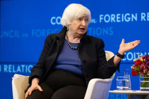 Janet Yellen, uma mulher idosa de cabelos brancos curtos com franja, está sentada em uma poltrona branca falando em um evento. Ela veste uma blusa azul-bic, um blazer escuro texturizado com a gola levantada, calça escura e um colar de contas pretas e prateadas. Ela está gesticulando com a mão esquerda aberta para o lado enquanto fala. Ao fundo, há um painel azul com a inscrição "Council on Foreign Relations" repetida em letras brancas. Em primeiro plano, no canto inferior direito, há uma pequena mesa redonda com dois copos de água e um arranjo de flores vermelhas e roxas.