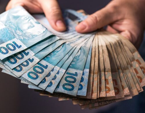 Pessoa segura e conta notas de R$ 100, representando restituição do Imposto de Renda e pagamento ao contribuinte.