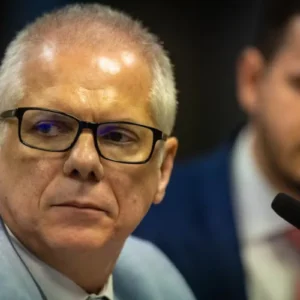 Foto em plano médio e de perfil de três quartos de um homem de pele clara e cabelos grisalhos bem curtos. Ele usa óculos de grau com armação preta retangular e veste um paletó cinza claro sobre camisa branca. O homem está com uma expressão séria, olhando ligeiramente para a esquerda. Ao fundo, fora de foco, aparece outro homem de terno escuro, camisa clara e gravata vermelha, além de um microfone de pedestal preto em primeiro plano à direita.