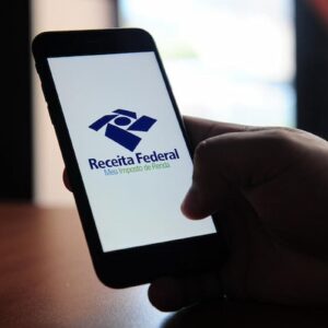Pessoa segura celular com aplicativo Meu Imposto de Renda da Receita Federal aberto na tela.