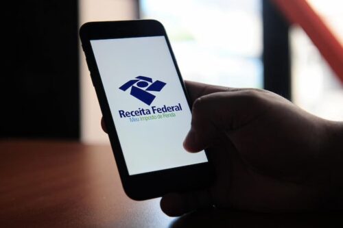 Pessoa segura celular com aplicativo Meu Imposto de Renda da Receita Federal aberto na tela.