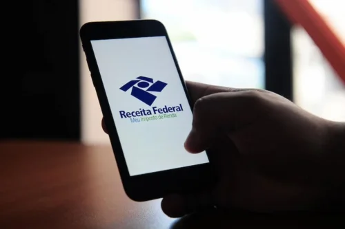 Pessoa segura celular com aplicativo Meu Imposto de Renda da Receita Federal aberto na tela.