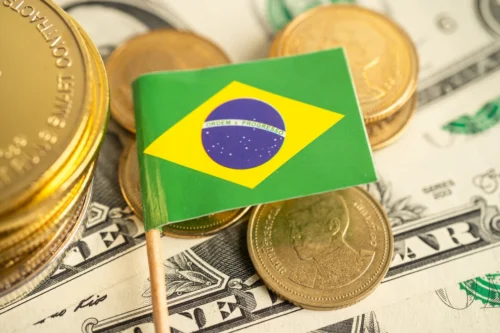 Bandeira do Brasil sobre moedas e notas de dólar, representando economia, inflação e investimentos.