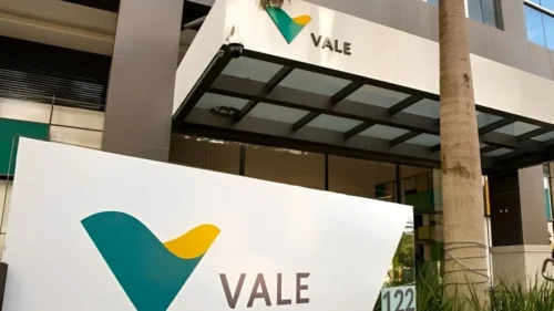 Placa com o logotipo verde e amarelo da Vale em primeiro plano e fachada de prédio corporativo ao fundo.